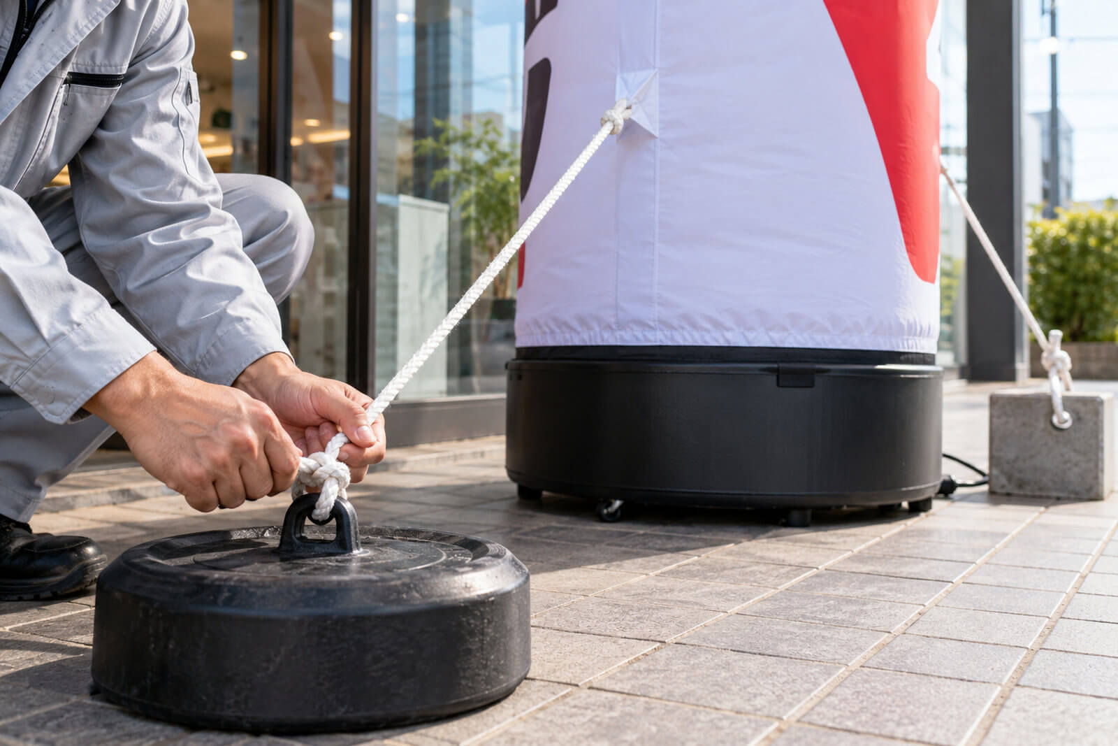 エアー看板のバルーン両側のタグにロープを通し、足元のウエイトとコンクリートブロックに結んで固定している手元