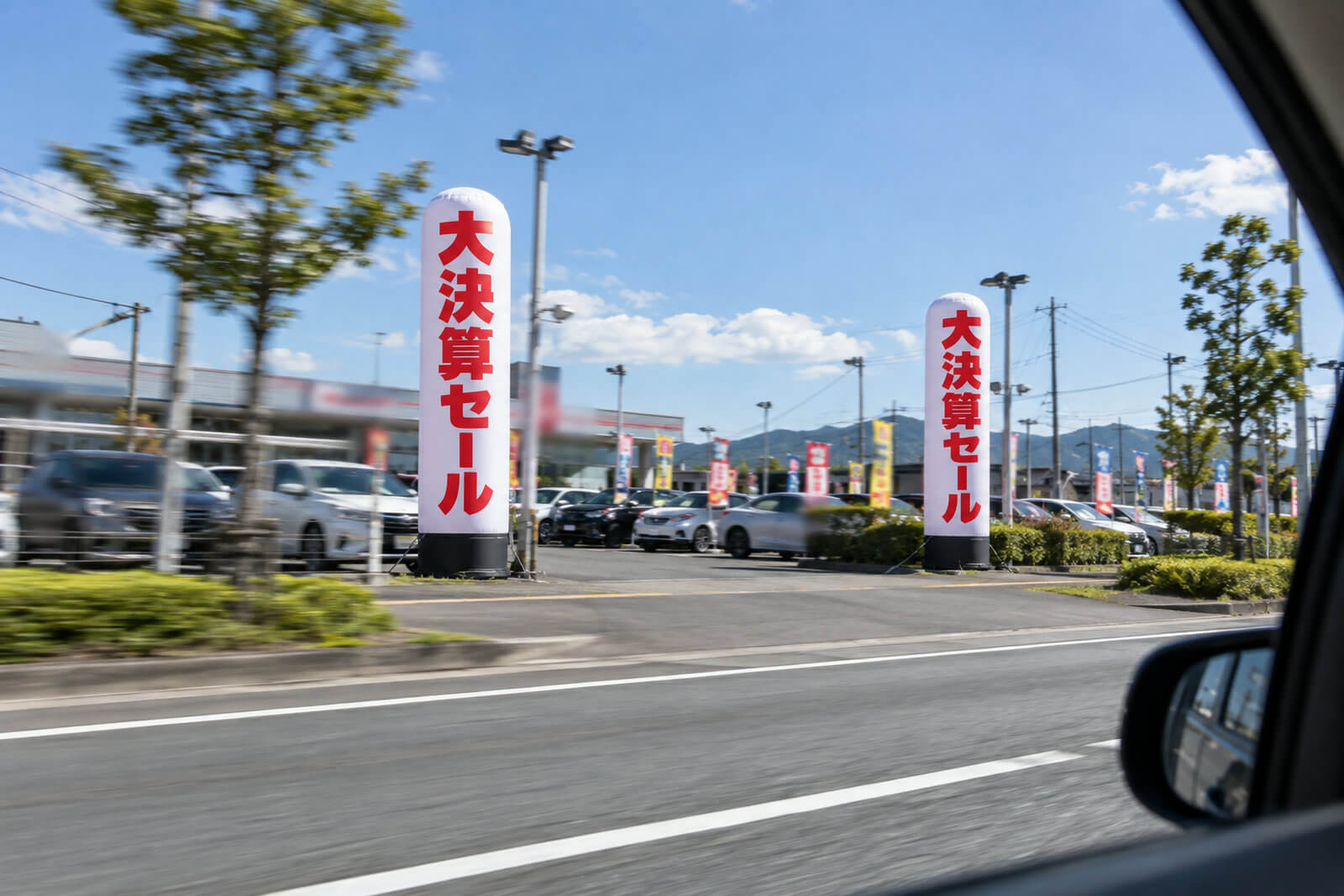 幹線道路沿いの中古車販売店の敷地に並んで立つ2台の高さ5mのエアー看板。走行中の車の窓越しに見える構図