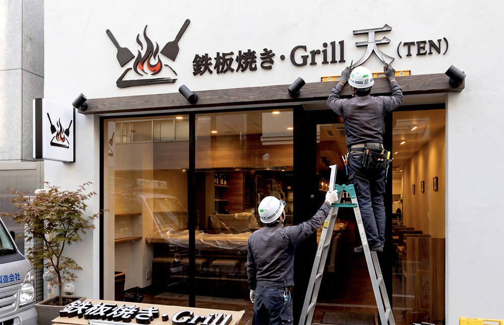 「Grill 天（TEN）Aoyama」様のSUS黒塗装チャンネル文字をファサードに取り付けている施工中の様子。職人2名が脚立で作業中。