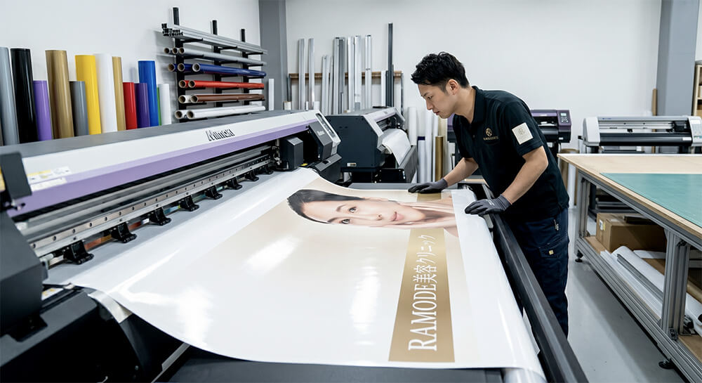 「RAMODE美容クリニック」様のウィンドウサイン製作工程。大型インクジェットプリンターで透明塩ビシートにシャンパンゴールドのグラフィックを印刷している様子。