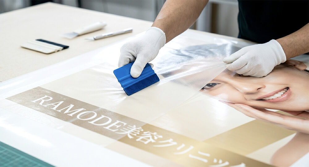 「RAMODE美容クリニック」様のウィンドウサイン製作工程。大判グラフィックシートにラミネートをスキージーで貼り合わせている職人の手元。