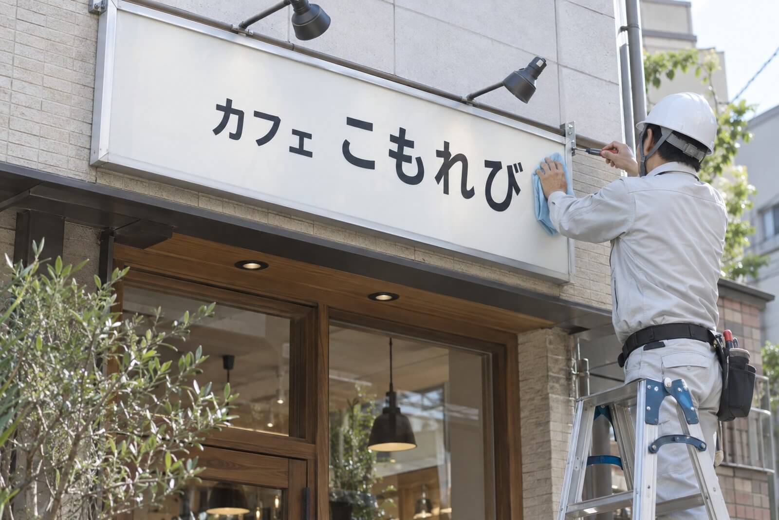 施工スタッフが脚立に乗って店舗のプレート看板のビスの緩みを確認し、表面を拭いている様子