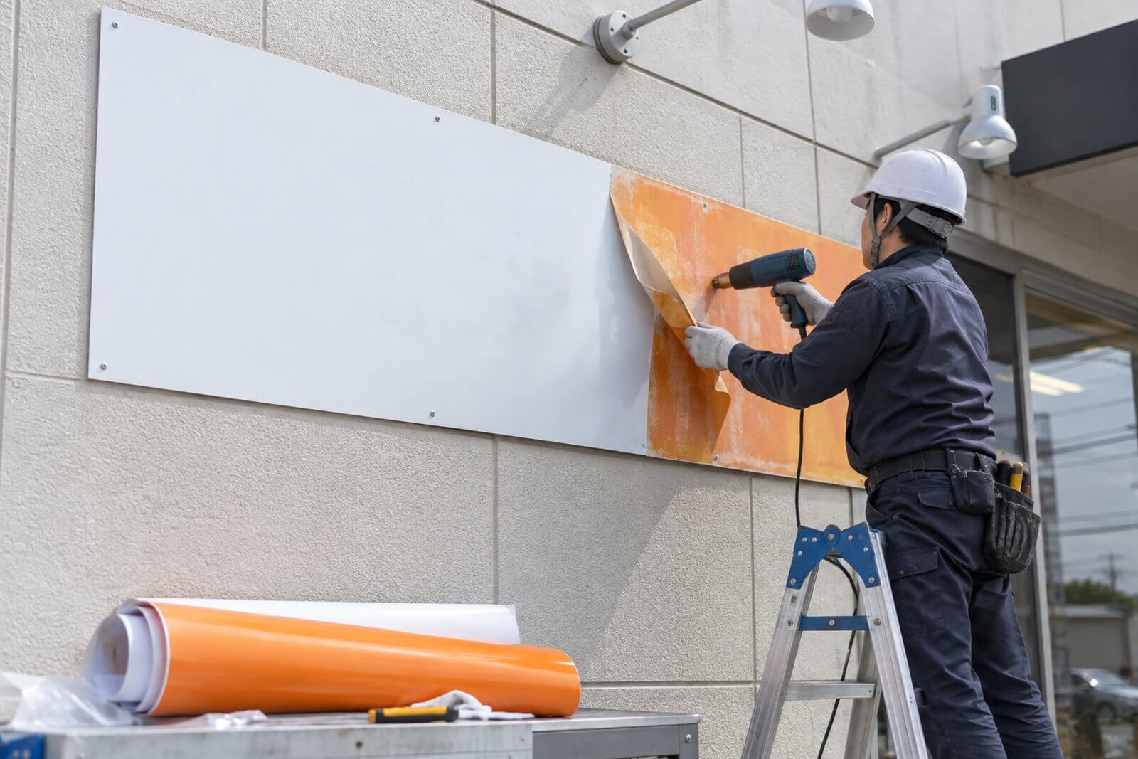 施工スタッフが壁に付いたままのプレート看板の古いシートをヒートガンで温めながら剥がし、新しいシートを貼っている様子