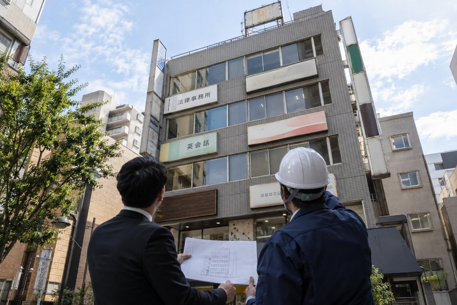 複数のテナント看板と袖看板が付いた中規模ビルを、管理会社の担当者と点検員が図面を見ながら見上げている