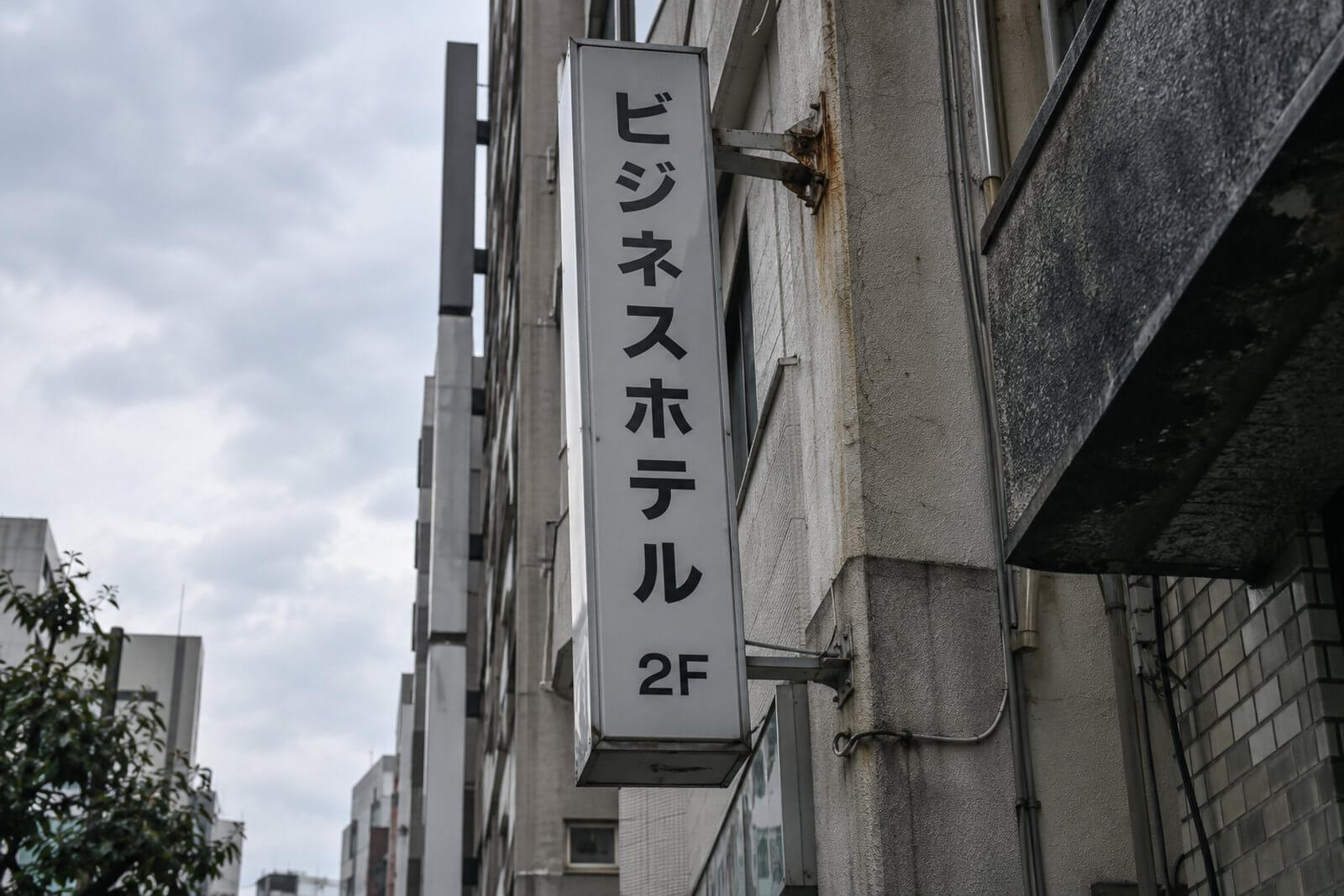 ビルの壁面から張り出した袖看板がわずかに傾き、支持アームの根元の塗装が剥がれている様子