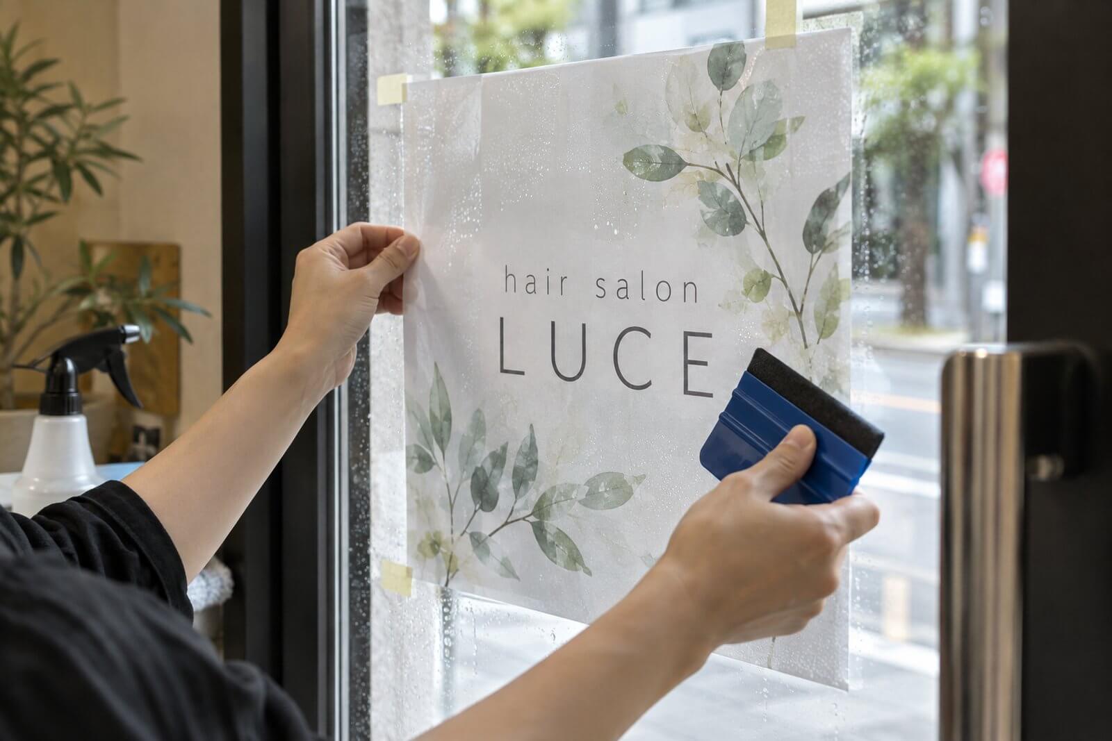 店舗のガラス扉に、霧吹きで水を吹きかけながらスキージでウィンドウサインのシートを貼っている手元
