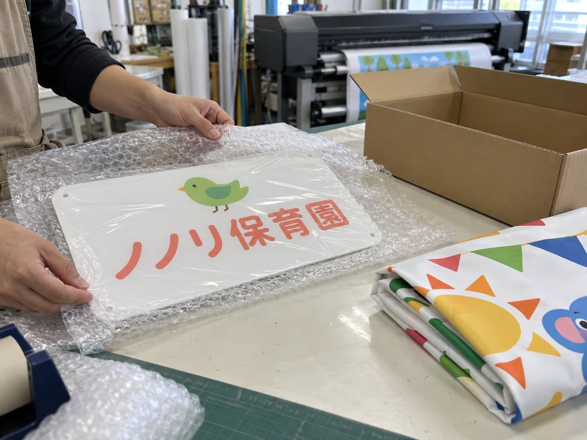 看板工場の作業台で、保育園の門の表示のアクリル看板と、運動会の幕を梱包している手元