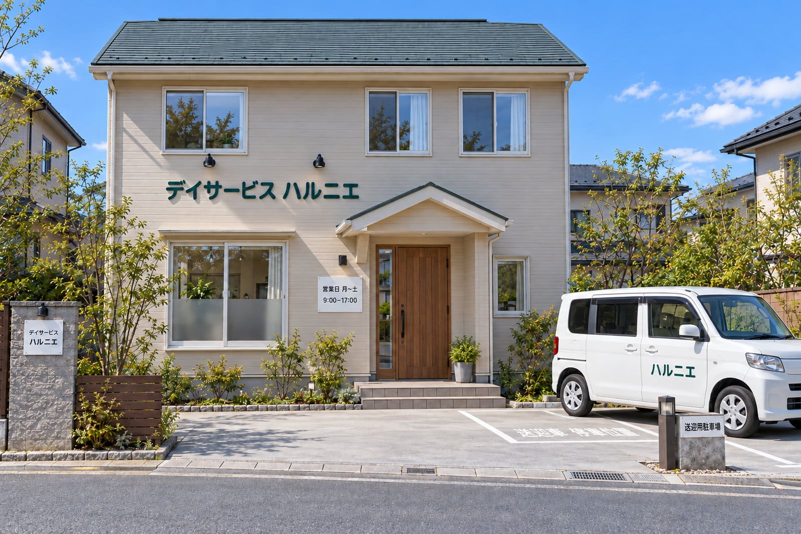 住宅街の2階建ての家を改修したデイサービスの外観。外壁にやわらかい緑色の立体文字の施設名、門柱に表札サイズの白い看板、玄関の横に営業日の表示、敷地の中に施設名を入れた白い送迎の車