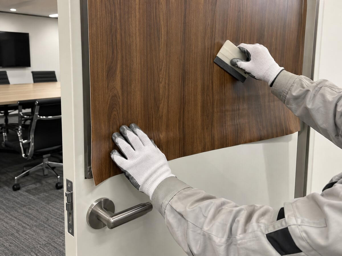 施工スタッフがオフィスのドアに木目調のダイノックシートをスキージーで空気を抜きながら貼っている手元