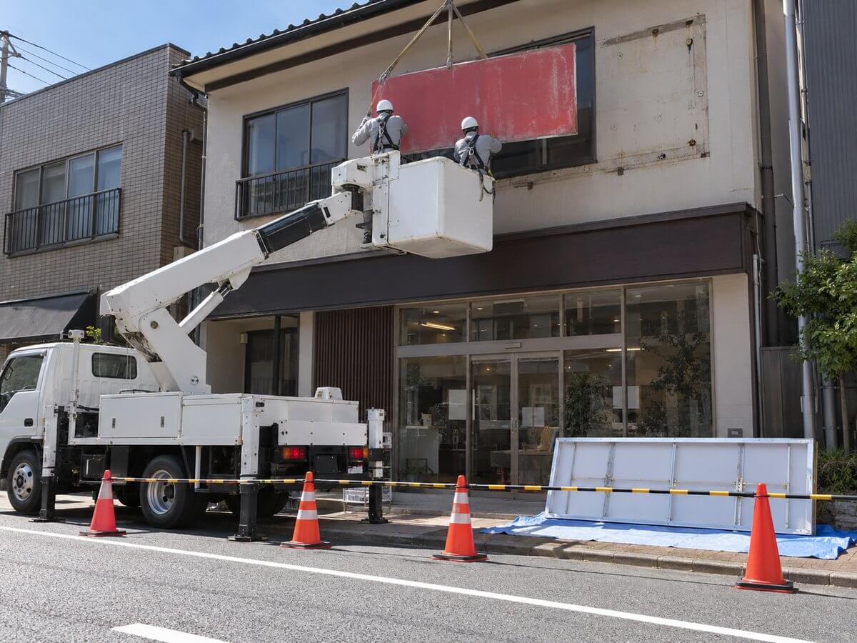 高所作業車に乗った施工スタッフが、店舗の外壁から古い看板を取り外し、足元に新しい看板が用意されている様子