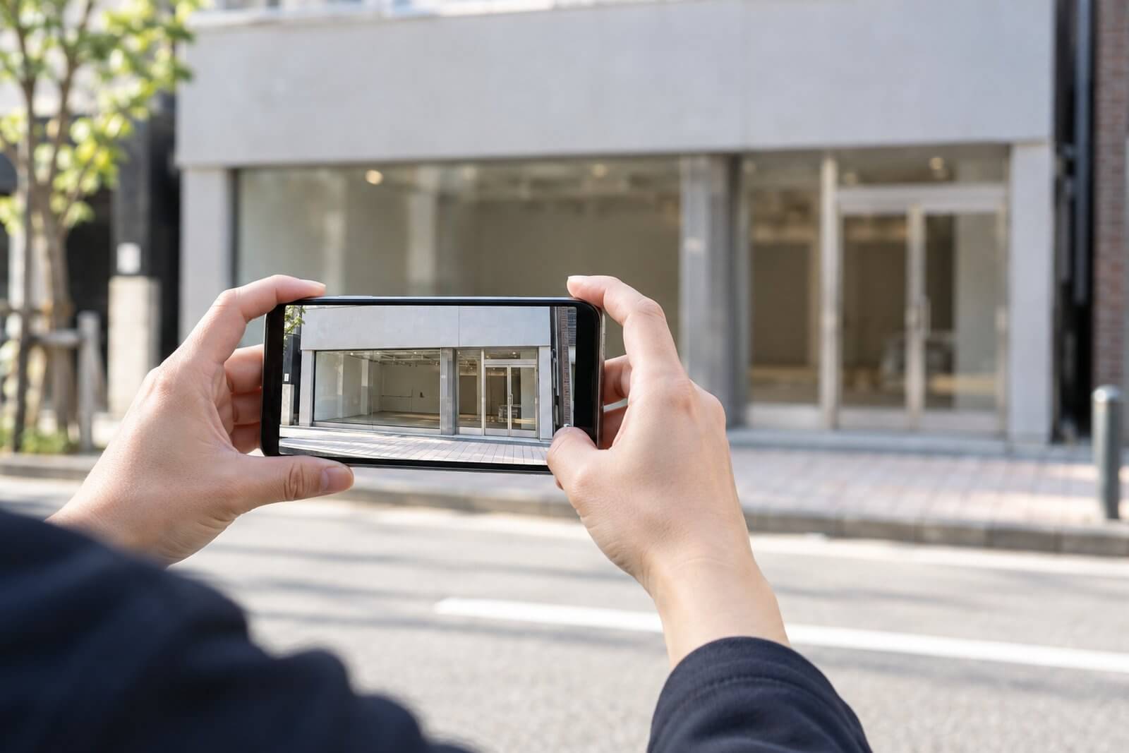 空き店舗の前の歩道で、スマートフォンを横向きに構えて建物の1階全体を撮影している人の手元