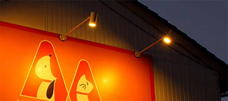 夕方、オレンジ色の店舗の看板を、看板の上に取り付けた2灯のアームスポットが電球色のあたたかい光で照らしている