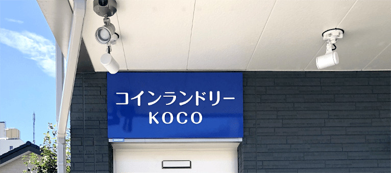 店舗の入口の軒下の天井に直付フランジで取り付けた白い本体のスポットライトが、青い看板を照らしている