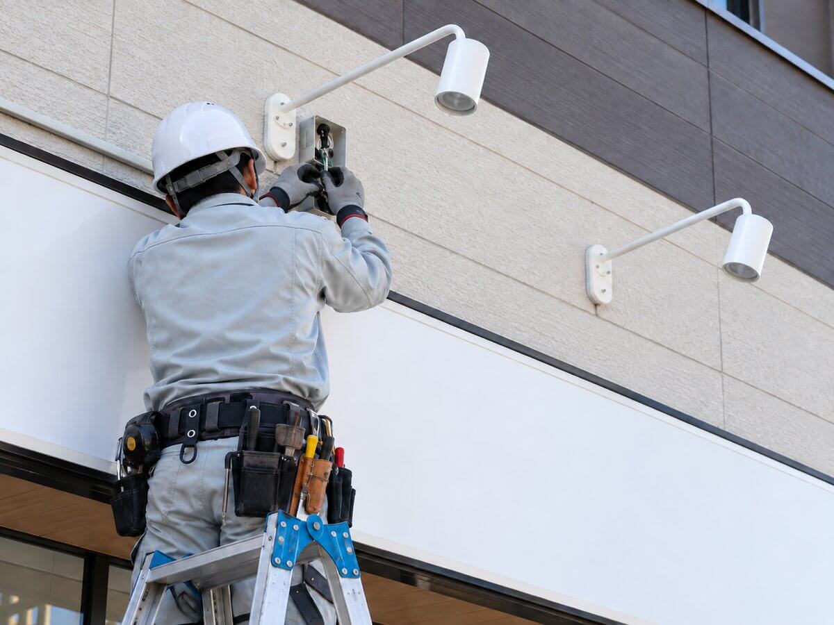 脚立に乗った作業員が、店舗の看板の上でアームスポットライトの配線をつないでいる手元