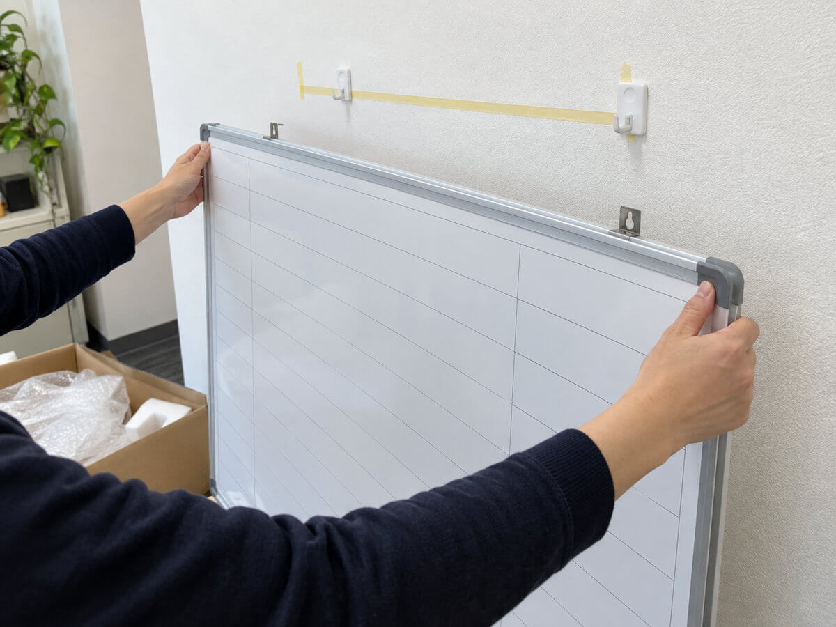 届いた特注ホワイトボードの箱を開け、壁に付けた2つのフックに吊り金具を合わせて掛けている手元。壁には水平を確かめるための紙のメモが貼られている