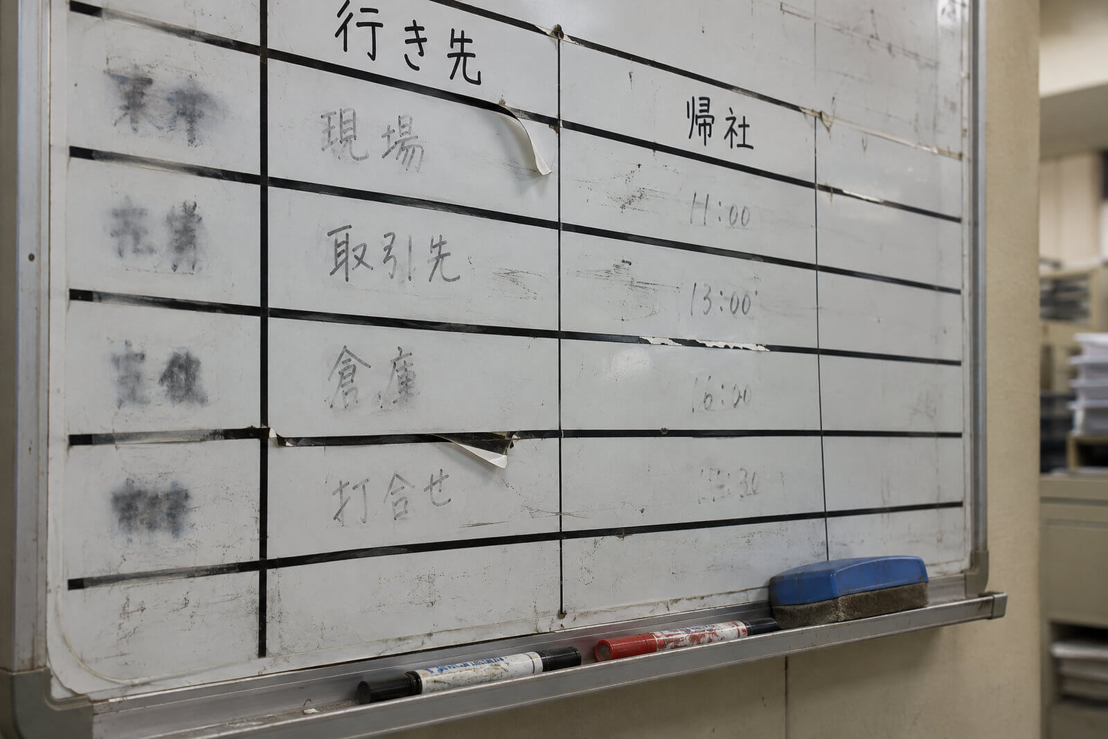 事務所の壁掛けホワイトボードのアップ。黒い線引きテープで作った行動予定表の罫線が端から剥がれ、消し残しのマーカーの跡が残っている